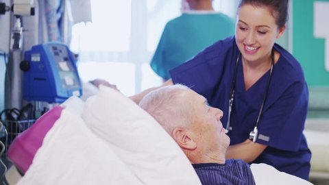 A beautiful female nurse attends to an elderly male patient, plumping up his pillows and chatting with him. In slow motion. 