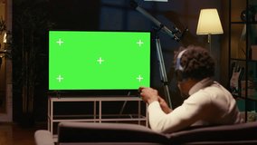 African american man sitting on couch at home, relaxing by playing videogames on green screen TV. Gamer unwinding in apartment by competing in online multiplayer game displayed on mockup television