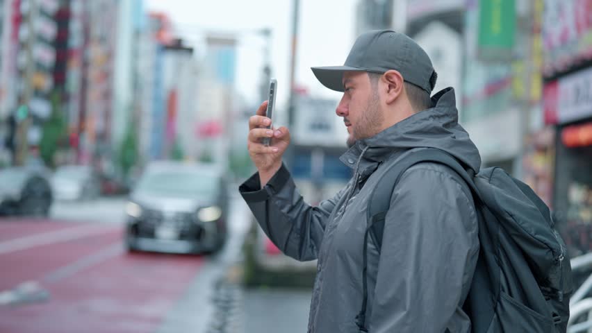 Side view of concentrated young Hispanic male tourist in warm clothes and cap with backpack taking photo on buildings on smartphone while standing in Akihabara neighborhood in Tokyo, Japan Royalty-Free Stock Footage #3421843471