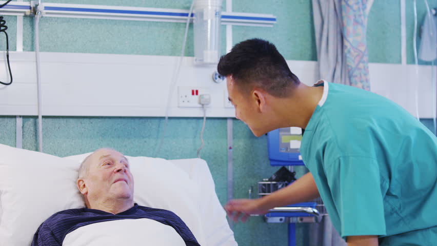 A male nurse attends to an elderly male patient, holding his hand and chatting with him.