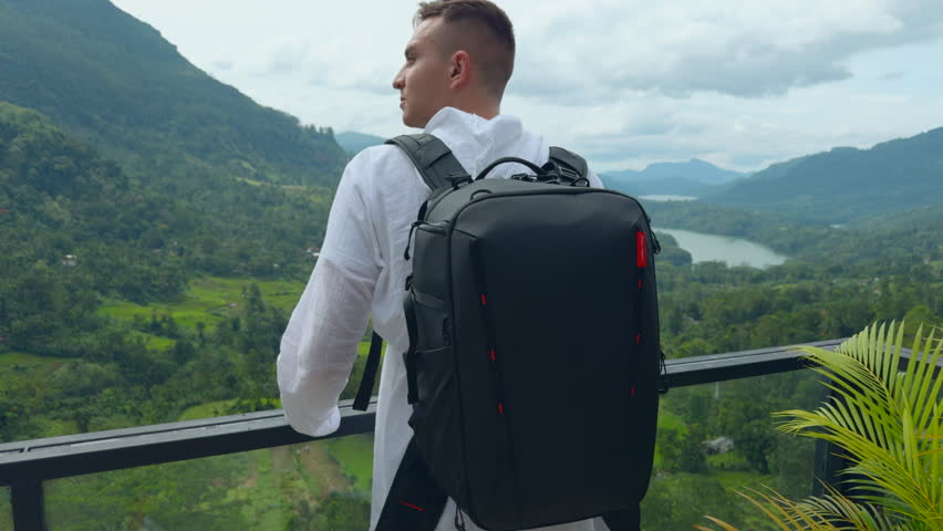 Man with view of tropical mountain valley. Action. Man on observation deck in tropical mountains. Man with backpack on sightseeing attraction overlooking green mountains