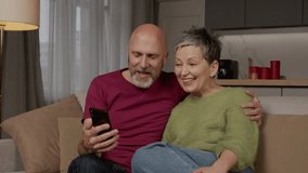 Joyful charming middle aged woman and excited bearded handsome man sitting on sofa, sharing cellphone and watching funny video content online while couple enjoying leisure in domestic room