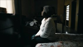 Pensive Senior African American woman in 80s seated by bedside with sad lonely look gazing at window in messy bedroom depicting solitude in old age - Powered by Shutterstock - Get 15% off with code: PIKWIZARD15