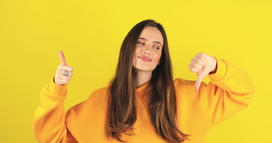 Young woman in yellow hoody making good-bad sign isolated on yellow background. Girl make choice, or makes a decision thumb up or thumb down, like or dislike, yes or no.