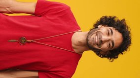 Vertical video, Happy attractive man with curly hair wearing red T-shirt, showing representing heart in shape of fingers gesture looking at camera isolated on yellow background in studio