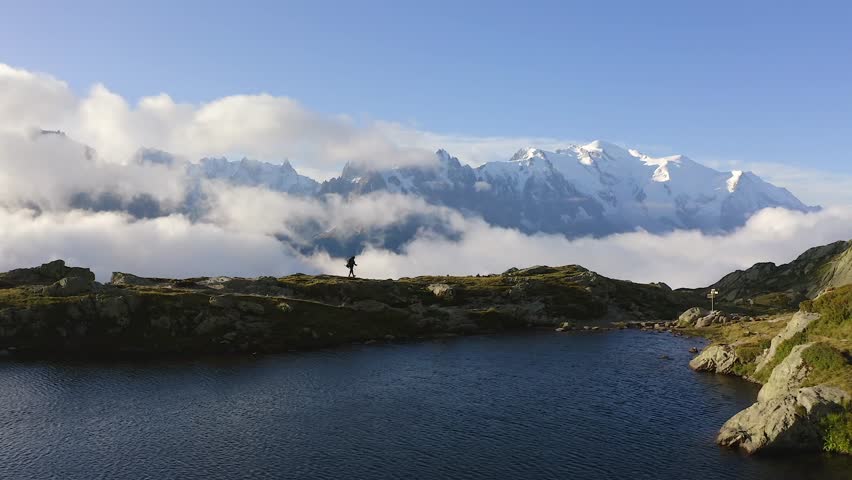 A drone captures a picturesque scene—an avid hiker strolling near a serene lake, while unveiling the grandeur of colossal snowy mountains towering in the background