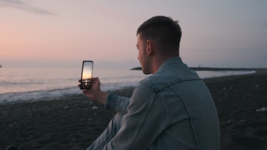 Young man tourist taking photo of sea sunset on black sand beach using smartphone. Male with mobile phone takes pictures, enjoys nature, freedom, relaxing. Traveler vacation concept Royalty-Free Stock Footage #3422396881