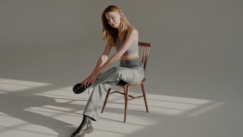 Young attractive woman in stylish jeans and top sitting on wooden chair and posing on camera during studio shooting. Beautiful smiling girl model