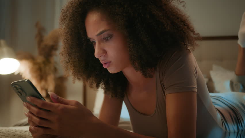Disappointed sad African American woman looking at mobile phone device upset frustrated ethnic female gen z girl lying on bed with smartphone worried scam message loss error failure at evening home Royalty-Free Stock Footage #3422497037