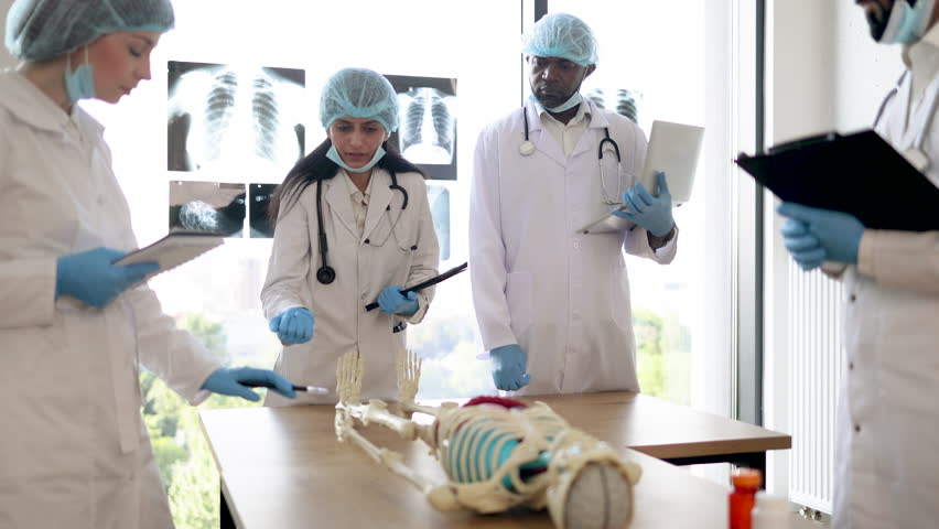 University, education, medicine, anatomy concept. Multiracial medical students in protective masks, gloves and hats with Indian female professor and human skeleton model in classroom, studying anatomy - Powered by Shutterstock - Get 15% off with code: PIKWIZARD15