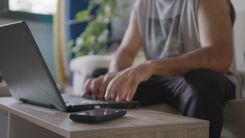 Young adult male working from home in his living room using laptop, distracted from work with a phone new mail pop up, sitting on the couch with casual clothes