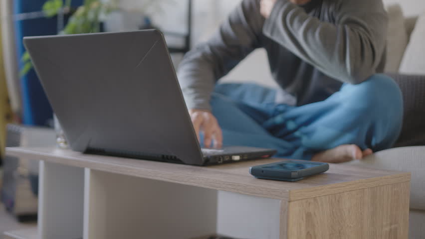 Young adult working from home with his pajamas, Uses the living room as an office together with a laptop, He is distracted by a reminder popup from his cell phone that says how to continue today Royalty-Free Stock Footage #3422530275
