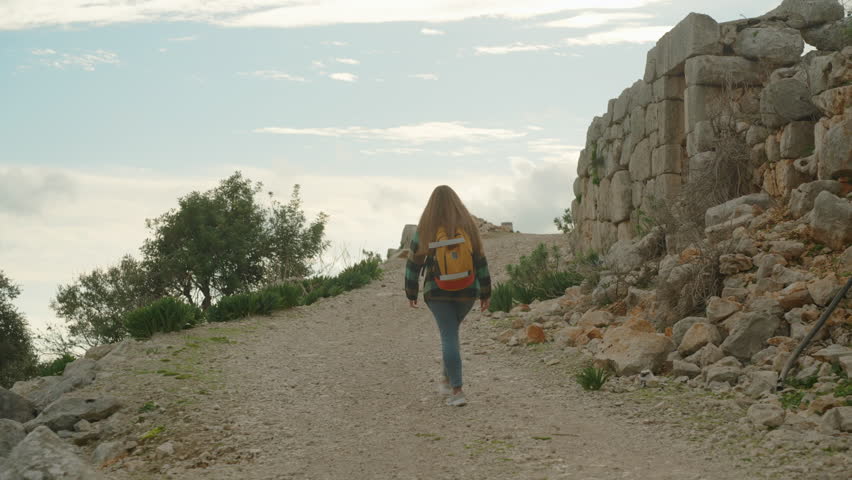 Following a young woman who ascends along a steep road to the mountain summit. Ancient ruins are on the edge near the cliff. Sunset casts warm light through the clouds.