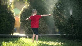 Slow motion video of funny little boy playing with garden sprinkler in sunny backyard. School child laughing, jumping and having fun with spray of water. Summer outdoors activity for children