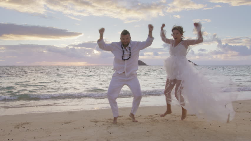 Crazy hilarious young couple dancing with raised arms on sandy tropical beach. Comic people celebrating wedding day on paradise island scenic nature. Happiness, freedom, excitement, love, joy concept