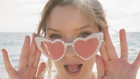 Close up young woman in pink glasses winking to camera on ocean background. Playful woman in glamour heart glasses looking to camera. Portrait fashion girl winking eye on beach. Fashion model wink eye - Powered by Shutterstock - Get 15% off with code: PIKWIZARD15