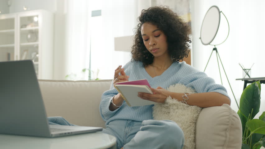 Focused biracial woman in casual blue clothes sitting of sofa. Young curly lady thinking through creative content writing 4K. Concentrated multi ethnic girl thinking and writing down plan in notebook