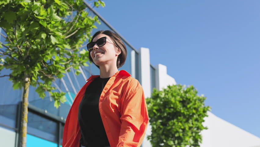 Low angle view of positive caucasian female dressed in casual attire and sunglasses walking throught sunny urban street. Attractive woman looking aside while moving near glassy building.