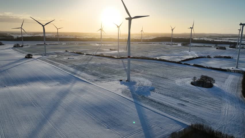 Aerial footage of a large spinning wind turbine within a wind farm in agricultural fields in winter. Winter snow on a rural agricultural field with view of wind turbine in the backlit. Royalty-Free Stock Footage #3422893077