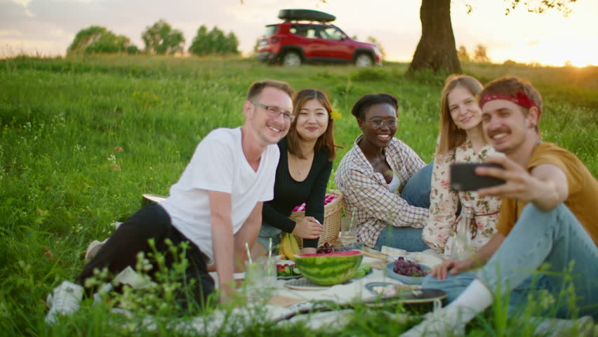 Happy Diverse group of people are taking Selfie by smartphone at picnic in park or stop on road trip. Young multiethnic friends have fun, eat and spend summer time together. 4K wide gimbal shot