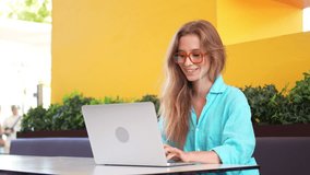 Smiling young female freelancer using laptop at cafe table. Beautiful businesswoman in blue shirt with long blond hair working remotely in coffee shop. Happy woman in trendy eyeglasses. - Powered by Shutterstock - Get 15% off with code: PIKWIZARD15
