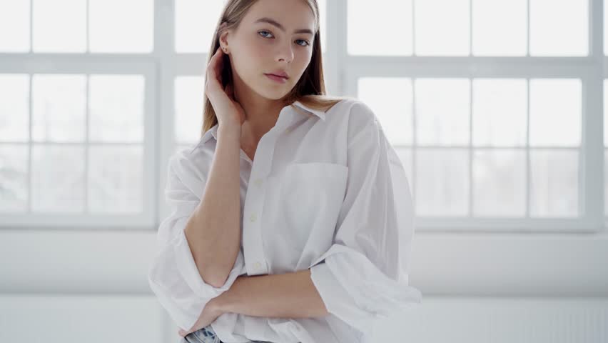 Confident woman in white shirt poses studio looking at camera. Woman poses symbol female beauty. Woman poses exemplifying modeling poise white Showcasing feminine beauty style