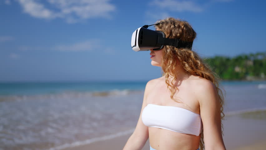 Woman explores virtual world on tropical beach in VR headset, sways to immersive experience in sunny setting. Natural landscape meets digital realm, blurring reality lines. Slow motion.