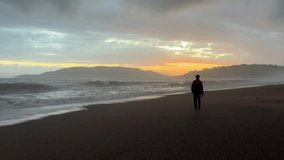 A man walks along a sandy beach along a stormy sea against the backdrop of a beautiful golden sunset.