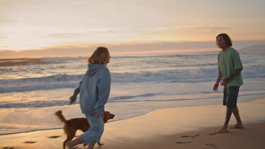 Carefree girl jumping guy hands at sundown shoreline. Young man carrying girlfriend in arms at sea beach. Two lovers walking dog having fun at summer ocean. Happy couple enjoying ocean nature together