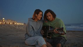 Smiling teenagers enjoying guitar music at evening sea zoom on. Relaxed musician man singing song touching strings closeup. Romantic couple resting on sand beach. Positive pair spending time together - Powered by Shutterstock - Get 15% off with code: PIKWIZARD15