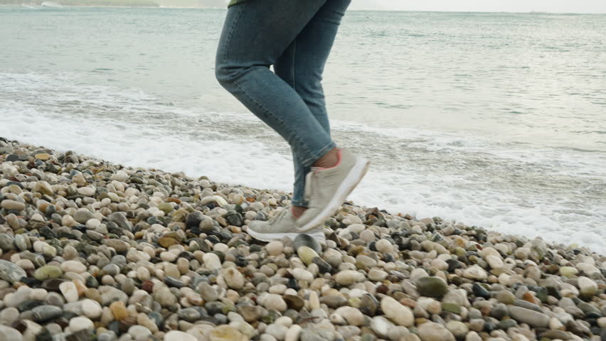A Woman in Sneakers and Jeans Walks Along a Pebble Beach, the Waves Almost Touching Her Feet. Royalty-Free Stock Footage #3423392759
