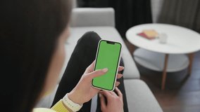 Young Brunette Woman Holding the Phone and Touching the Screen. Over Shoulder View of Hands of the Girl Resting on the Sofa Enjoys Spending Time with the Chroma Key Phone. Green Screen Concept