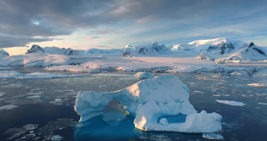 Sunset Antarctic iceberg floating in icy ocean. Snow-capped mountains in background. Fly over the untouched wilderness of Antarctica colorful landscape. Cinematic aerial drone panorama zoom out.