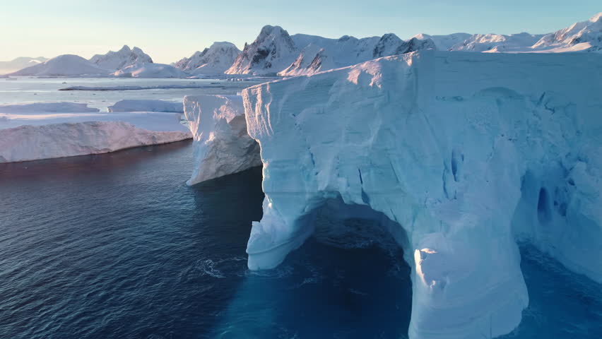 Antarctica huge iceberg with caves and arch in sunset light. A breathtaking scene of Antarctic big glacier in cold ocean waves, snow-capped mountains in background. Aerial panoramic drone footage