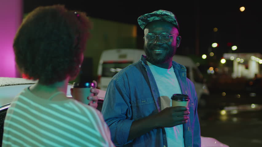 Over shoulder footage of stylish African American guy drinking coffee or tea and talking to unidentified Black lady standing near white car at neon lit fueling station at night Royalty-Free Stock Footage #3423815307