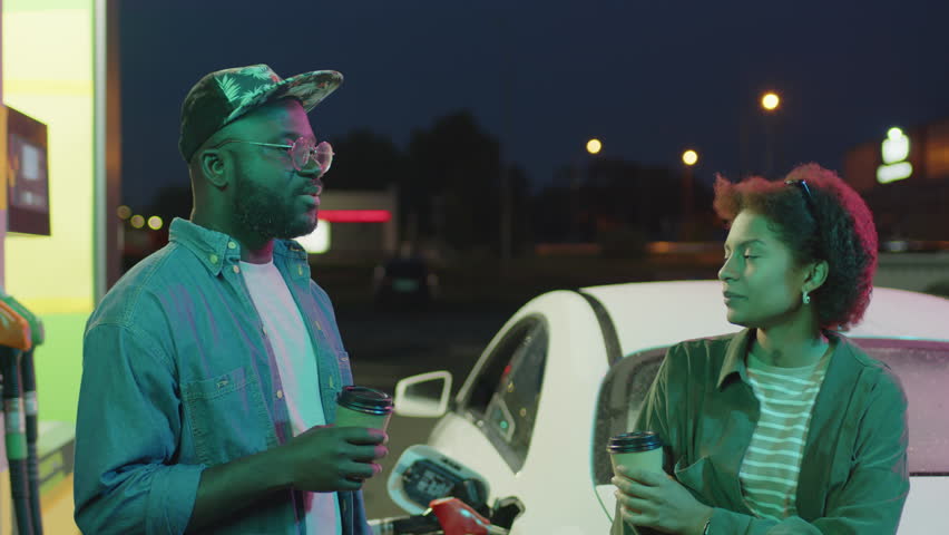 Medium footage of African American friends speaking near white modern car and drinking coffee or tea while waiting vehicle to refuel at neon lit petrol station at night Royalty-Free Stock Footage #3423872585