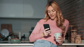 Video of pretty young woman using her mobile phone while drinking coffee in the kitchen at home.