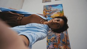 Medium vertical selfie shot of young African American woman shooting video on smartphone for social media while visiting contemporary art exhibition in museum, and showing paintings to followers