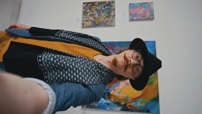 Medium vertical selfie shot of eccentric Caucasian male artist shooting video on smartphone at personal exhibition in art gallery, in front of abstract paintings, and urging subscribers to visit it
