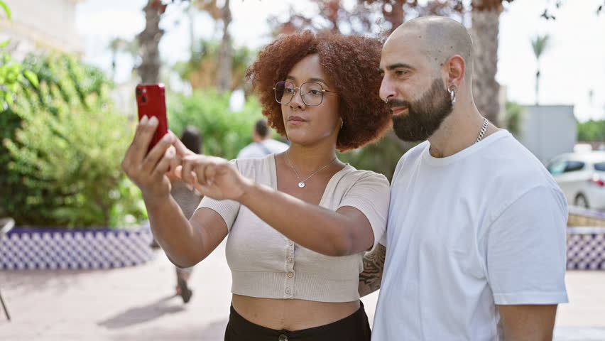 Beautiful couple smiling, standing together in park, enjoying outdoor selfie fun with smartphone