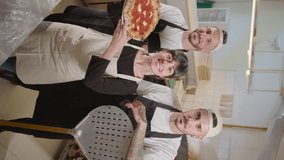 Team of pizzeria workers in aprons posing together on camera in the kitchen. Beautiful female baker holding just cooked pizza and smiling as two male colleagues standing behind. Vertical clip