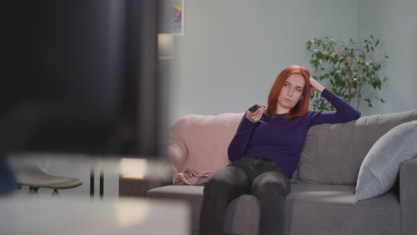 Overworked housewife sits on couch changing channels on TV with remote control. Red-haired woman spends evening in living room at home after work Royalty-Free Stock Footage #3424170615
