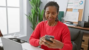 Smiling african american woman using smartphone in modern office with plants and laptop.
