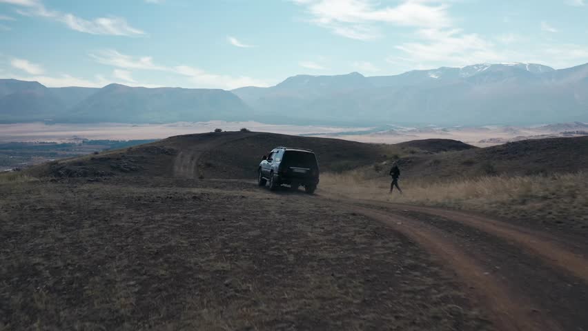 Black off-road SUV automobile driving on dirt dust grail road up to hill at natural meadow with mountains rock drone follow shot. Auto trip steppe travel journey adventure freedom. Man walking to car