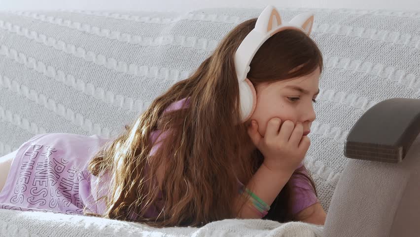 A happy little girl in headphones with cat ears listens to music lying on a gray sofa in the living room, close-up.