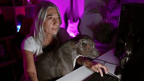 Grey-haired middle age streamer woman and pet dog dive into the world of cyber gaming, playing intense video game in home gaming room during a late-night streaming session.
