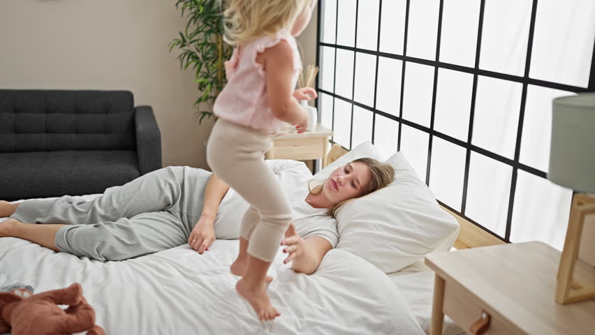 Caucasian mother and daughter bonding, jumping on the bed together, smiling inside their cozy bedroom, waking up a happy home with joyful expressions. Royalty-Free Stock Footage #3424264903