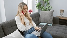 A concentrated young woman using a laptop and talking on a phone in a modern living room.