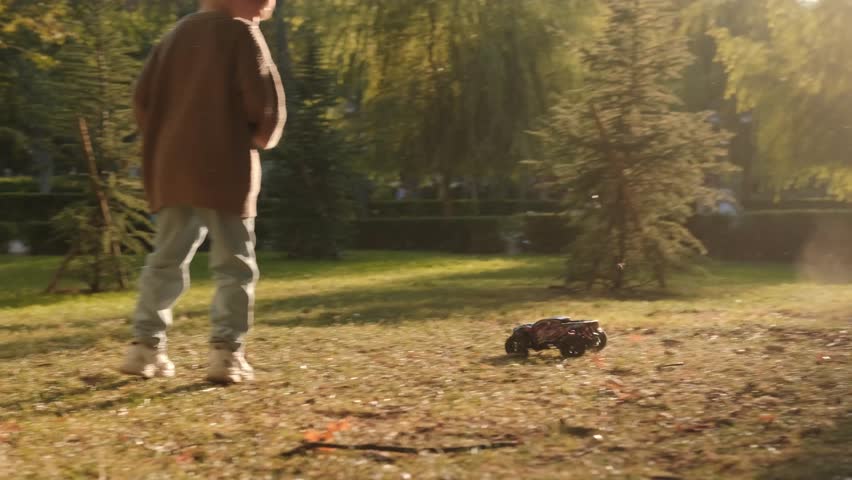 A boy stands in a sunlit clearing in a park and plays with a remote control car that moves quickly through the grass around him. Outdoor games, radio controlled toys, childhood. Royalty-Free Stock Footage #3424294165