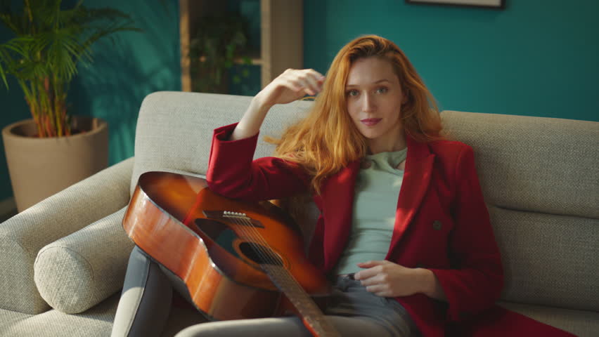 Handheld shot of a young beautiful girl smiling and looking into the camera with a guitar in her lap, she is sitting on the sofa